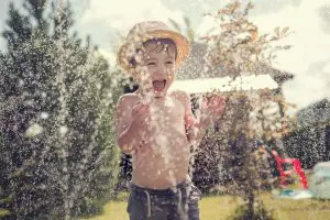 Cute little boy in straw hat is laughing and having fun running under water spraying hose.